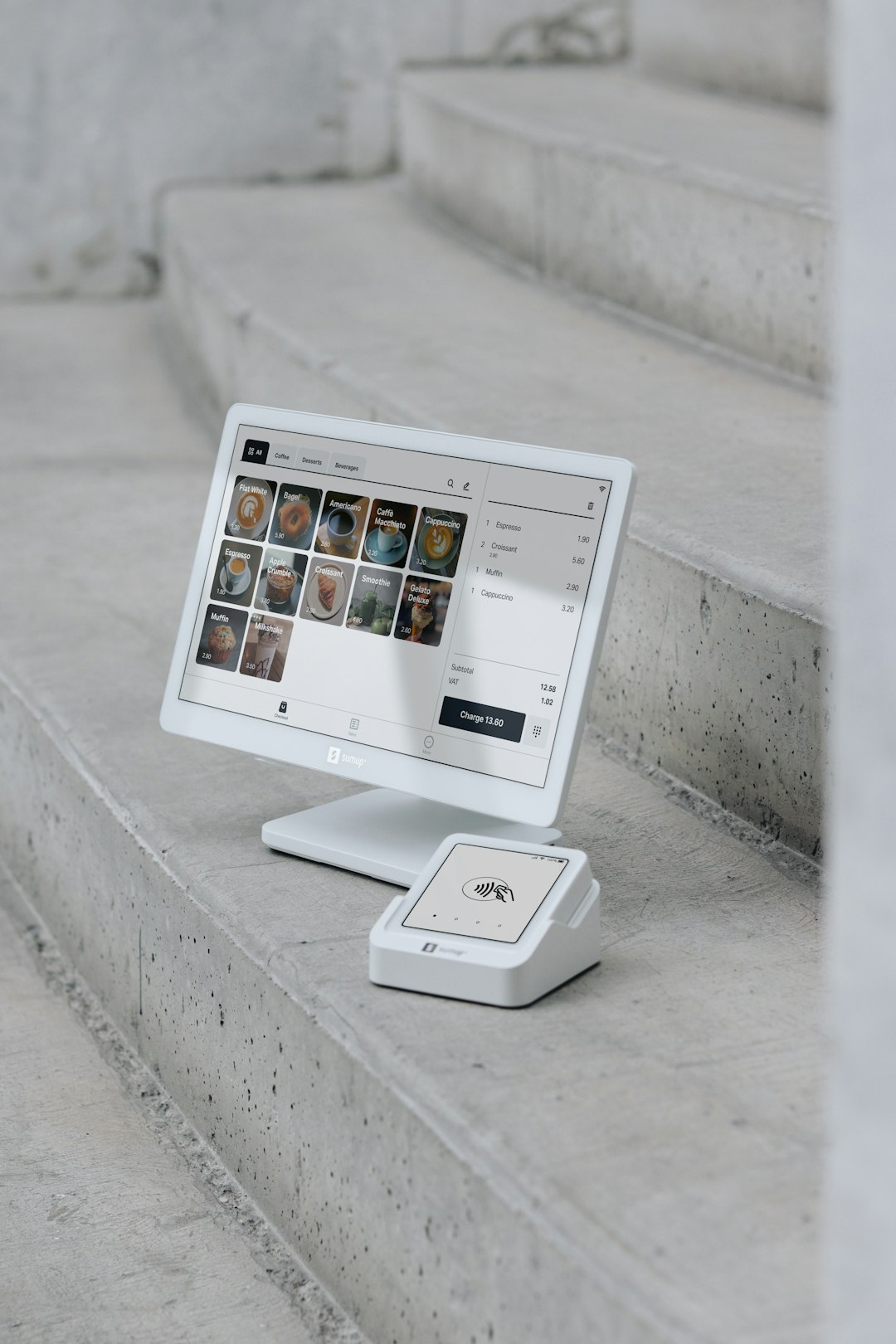 A point of sale (POS) setup is displayed on concrete steps, featuring a modern white display screen alongside a compact card reader. The screen shows a digital menu with various products, while the card reader is ready for contactless payments. This clean, minimalistic setup highlights the portability and sleek design of SumUp’s payment solutions, ideal for small businesses, cafes, and retail environments that value both functionality and aesthetics.